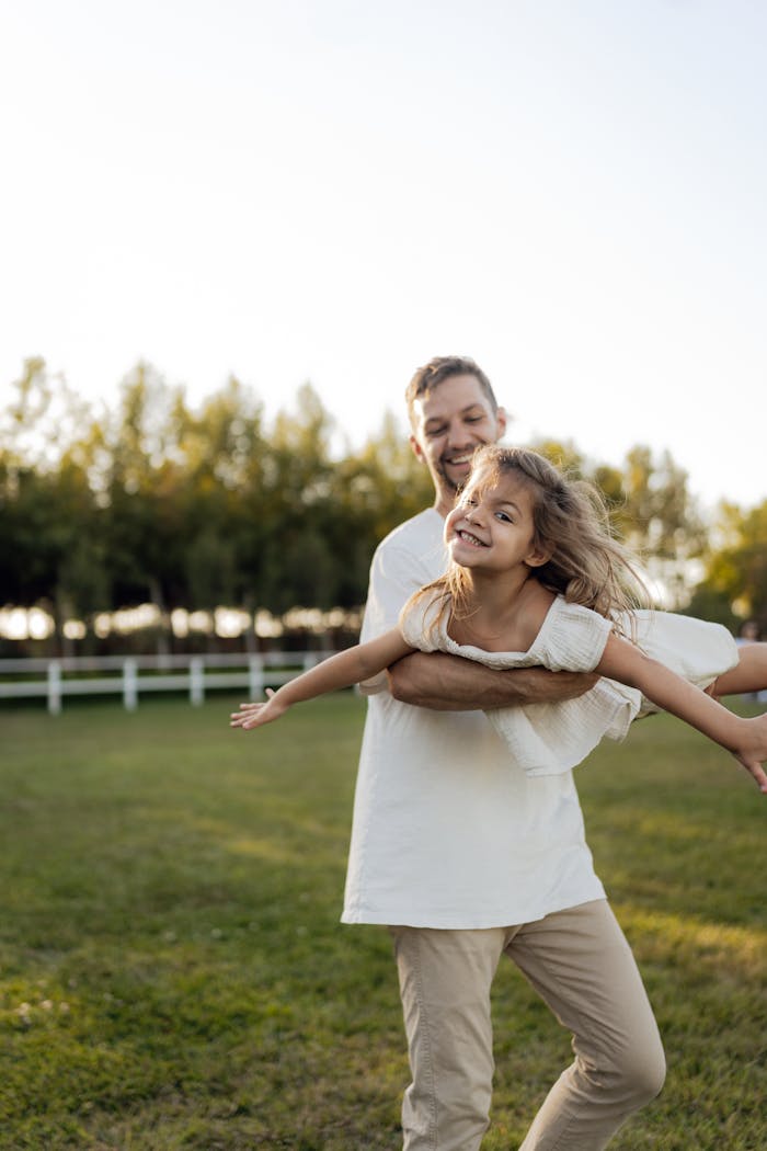 A cheerful father and daughter playfully pose in a green park, surrounded by trees and sunshine.