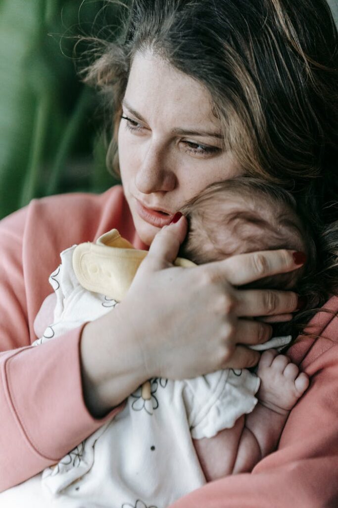 Upset mother with little newborn baby in arms looking into distance with serious gaze while standing in light room at home